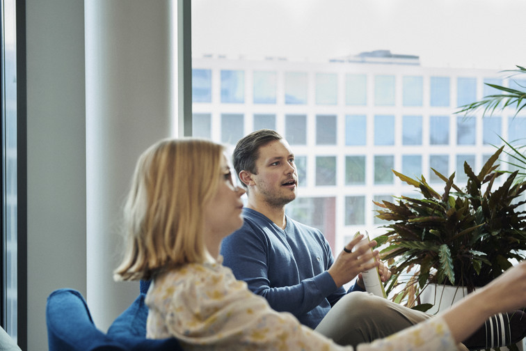 Two colleagues sit in a cheery office and speak with a third person, not visible.