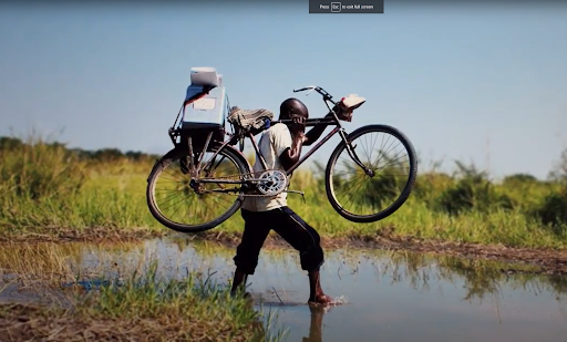 A man carries his bike with a cooler containing the Covid vaccine across a river.